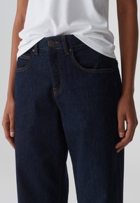 Jeans in denim blu scuro con chiusura a bottone, cinque tasche e cuciture dorate; il tessuto presenta una leggera texture e una vestibilità classica.