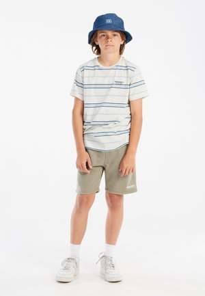 T-shirt a righe a maniche corte in beige e blu, abbinato a pantaloni corti verde oliva. Sneakers bianche e un cappello a secchiello blu completano l'outfit.