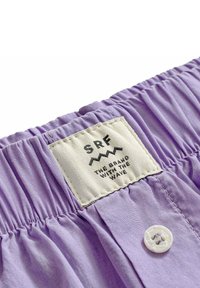 Close-up of a lavender garment with elastic waistband, a white button, and a white sewn label reading "SRF The Brand With The Wave."