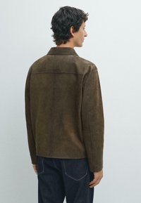 Brun mockajacka med krage, rak klippning, fickdetaljer och en mjuk textur, bärs över mörka denimbyxor.