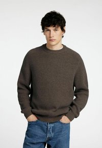 Bruine geribbelde gebreide trui met een ronde halslijn, lange mouwen en een relaxte pasvorm, gecombineerd met blauwe denim jeans.