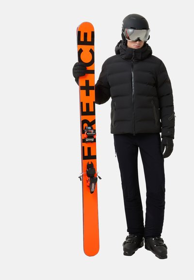 Osoba ve černém lyžařském vybavení a helmě drží vertikálně jasně oranžovou lyži s nápisem "FREE+RICE" na jednobarevném pozadí.