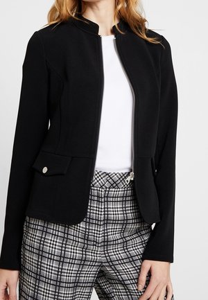 Vrouw draagt een nauwsluitend zwart jasje met ritssluiting aan de voorkant en klepzakken, een wit topje en hooggetailleerde zwart-wit geruite broek.
