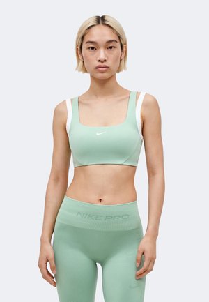 Γυναίκα με κοντά ξανθά μαλλιά, φορώντας αθλητικό μπουστάκι Nike σε μέντα και ψηλόμεσα κολάν, που στέκεται μπροστά σε απλό φωτεινό φόντο.