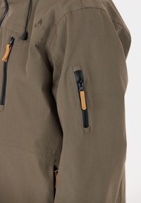 Ruskea kangastakki, jossa on vetoketjulla varustettu tasku hihaisessa, kontrastivärinen musta vetoketju ja nahasta tehty vetotäppä helppoa käyttöä varten.