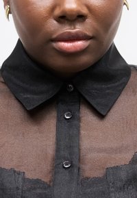 Κοντινό πλάνο ενός ατόμου που φοράει διάφανη μαύρη μπλούζα με σταθερό μαύρο γιακά και κουμπιά, εμφανίζοντας τα χείλη, το κάτω μέρος της μύτης και ένα χρυσό σκουλαρίκι.