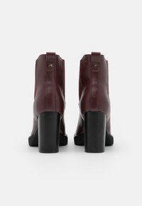 Burgundowe sztuczne skórzane botki na obcasie z kwadratowym noskiem, teksturowana czarna podeszwa i złoty detal z logo na pięcie, widok z tyłu.