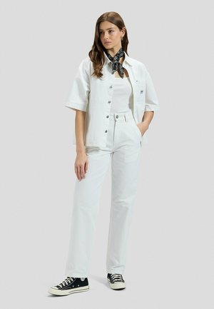 Femme portant une tenue entièrement blanche avec une chemise courte boutonnée, un pantalon taille haute, un foulard noir à motifs et des baskets noires, debout devant un fond gris.