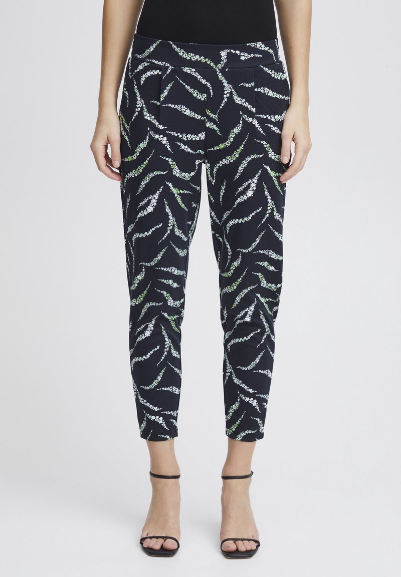 Femme portant un pantalon bleu marine longueur cheville à imprimé feuille abstrait blanc et vert, un haut noir sans manches et des sandales à talons noires.