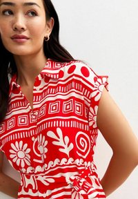 Camisa de estampado floral rojo con motivos blancos, con botones en el frente, mangas cortas, cuello y un detalle de cintura anudada. Textura de tela suave.
