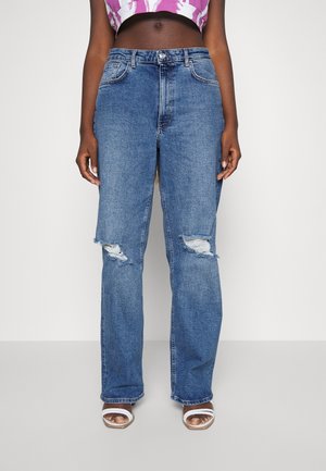 Kvinde iført blå højtaljede slidte jeans med huller ved knæene, en lilla og hvid cropped top og hvide sandaler med åben tå.