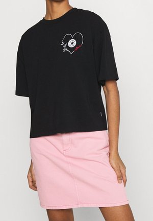 Schwarzes, kurzes T-Shirt mit kurzen Ärmeln, das ein weißes Herzdesign und pinke Nähte zeigt. Kombiniert mit einem hellrosa Jeansrock.