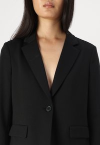 Blazer nero su misura con revers, chiusura a un bottone nero e due tasche frontali; texture liscia, design aderente.