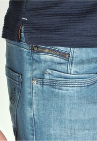 Light blue denim jeans with a visible zipper pocket, copper hardware accents, and a patch pocket on the side. Textured navy top.