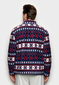 Uomo che indossa una giacca in pile a motivi navy, rossa e bianca con disegni di fiocchi di neve, in piedi di spalle contro uno sfondo neutro.