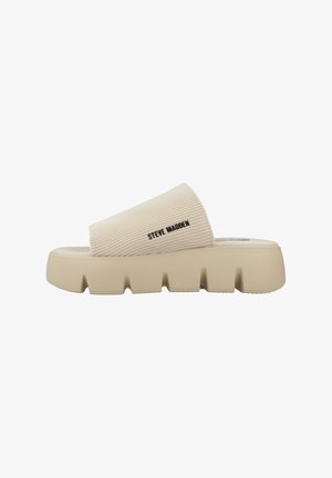 Sandalia de slide beige con parte superior de tela acanalada, suela de goma gruesa y diseño con cortes. Logo negro de "Steve Madden" mostrado.