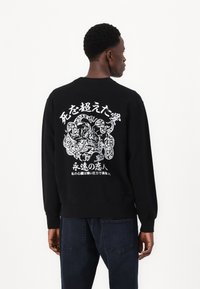 Sort sweatshirt med lange ærmer, der har et hvidt grafisk design på ryggen, som viser en drage og tekst på japansk.
