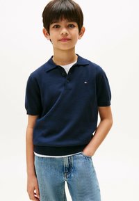 Granatowa koszulka polo z krótkim rękawem, z trzema guzikami oraz logo na piersi, zestawiona z jasnoniebieskimi dżinsami.