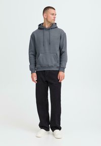 Joven mirando hacia la izquierda, vistiendo una sudadera gris, jeans negros y zapatillas blancas, contra un fondo claro y liso.