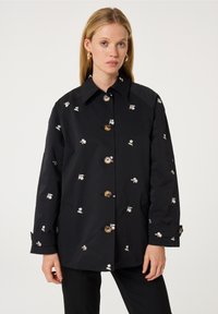 Zwarte jas met bloemmotief, grote knopen en een klassieke kraag. Beschikt over lange mouwen en zijvakken. Soepele textuur.