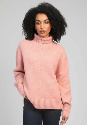 Femme aux cheveux bouclés portant un pull à col roulé rose pâle et un jean foncé, debout devant un fond gris clair uni.