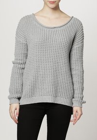 Femme portant un pull en tricot texturé gris clair à manches longues et un pantalon noir sur un fond uni.