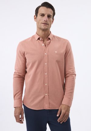 Mand iført en knappet salmonfarvet langærmet skjorte med en lille fuglelogo på brystet, kombineret med mørke bukser.