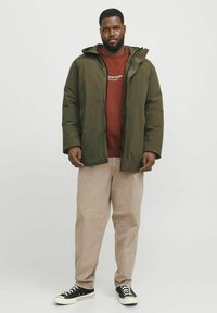 Green insulated jacket with a hood, zip and snap closure, worn over a red sweatshirt. Paired with light beige pants and black sneakers.