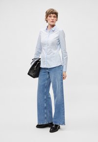 Svetlomodrá košeľa na gombíky v kombinácii s modrými džínami s širokými nohavicami a čiernymi koženými topánkami. V ľavej ruke držená čierna kabelka.