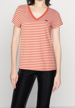 Kobieta ubrana w pomarańczowo-biały prążkowany t-shirt z dekoltem w serek z krótkim rękawem oraz czarne teksturowane spodnie, stojąca na jednolitym tle.