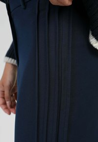 Tissu de pantalon tailleur bleu marine présentant des plis verticaux marqués, une texture lisse, et un gros plan d'une main près de la taille.