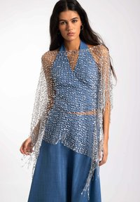 Un chal de malla plateada se posa sobre un top halter azul y una falda a juego. La tela texturizada presenta una trama suelta y abierta y bordes desflecados.