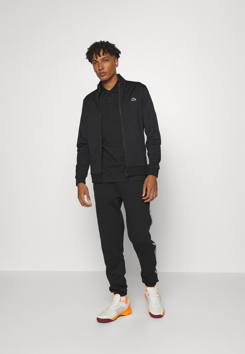 Zwarte zip-up jas over een zwart poloshirt, gecombineerd met zwarte joggers met zijkanten, en witte schoenen met oranje accenten.