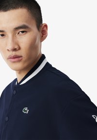 Chaqueta tipo varsity de color azul marino con un cuello de canalé estriado blanco, cierre de botones y un pequeño logotipo bordado en el pecho.