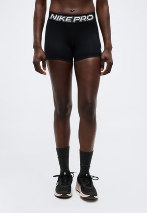 Atleet draagt zwarte Nike Pro shorts, donkere sportsokken en zwarte Nike hardloopschoenen, staand tegen een effen lichte achtergrond.