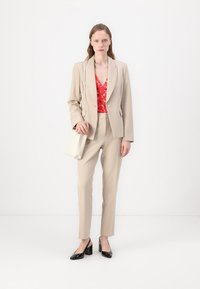 Beżowy garnitur o slim-fit z marynarką na jeden guzik, czerwoną bluzką w kwiaty, spodniami wąskimi, czarnymi szpilkami i kremową torebką. Gładka tekstura całości.