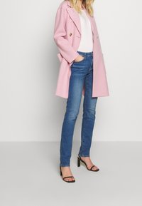 Eine Frau trägt einen hellrosa Mantel, ein weißes Oberteil, blaue Jeans und schwarze hochhackige Sandalen und steht vor einem schlichten hellen Hintergrund.