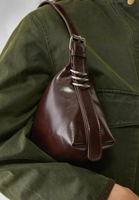 Borsa a tracolla in pelle marrone a forma triangolare, con accessori in argento e una tracolla. Sullo sfondo si vede una giacca verde texturizzata.