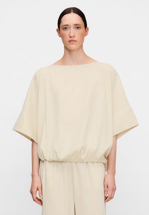 Frau mit glattem dunklem Haar trägt locker sitzendes, cremefarbenes Oberteil mit weiten Ärmeln und geraffter Taille, steht vor neutralem Hintergrund.