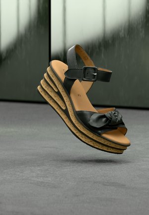 Black leather wedge sandal with knotted detail, adjustable strap, cork-textured sole, and layered design. Features open toe and padded insole.