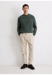 Pulover din tricot ribat de culoare verde închis, pantaloni din corduroy bej și pantofi negri; stând împotriva unui fundal texturat deschis.