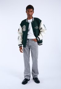 Chaqueta varsity verde y crema con parches bordados, sobre una camiseta blanca. Combinada con jeans gris claro y zapatos de plataforma negros.