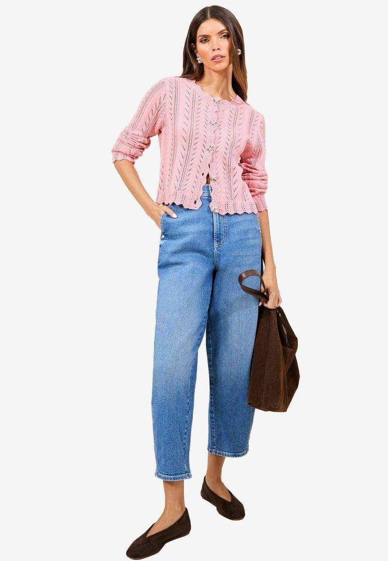 Rosa gestrickte Strickjacke mit Wellenkante und dekorativen Knöpfen, kombiniert mit hochgeschnittenen blauen Denim-Jeans und braunen Wildleder-Flats.