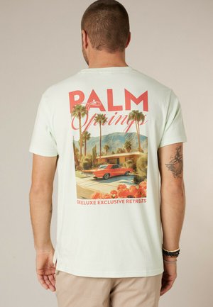 Man draagt lichtgekleurde t-shirt met afbeelding van een rood vintage auto, palmbomen en een huis uit het midden van de eeuw, tekst "Palm Springs" en "Deluxe Exclusive Retreats."