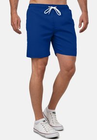 Blauwe zwembroek voor heren met een elastische tailleband, witte trekkoord en zijzakken. Vlotte textuur en knielange pasvorm. Witte sneakers.