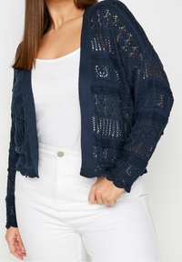 Marineblauer Strickcardigan mit offenem Gitterdesign, langen Ärmeln und gewellten Kanten, getragen über einem weißen Oberteil und kombiniert mit weißen Hosen.