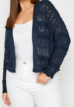 Mørkeblå strikket cardigan med åben gitterdesign, lange ærmer, scallopkanter, båret over en hvid top og parret med hvide bukser.
