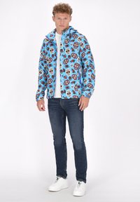 Blå jakke med et mønster af farverige kranier og blomster, parret med en hvid t-shirt, mørke jeans og hvide sneakers.