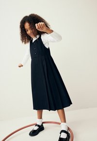 Marineblaue Latzkleid mit einem plissierten Rock, ausgestattet mit einem geknöpften Oberteil und einem weißen Langarmhemd. Getragen mit schwarzen Schuhen und Knöchelstrümpfen.
