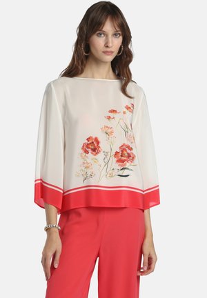 Femme portant un chemisier blanc avec des imprimés floraux rouges et des poignets rouges, assorti à un pantalon rouge, accessoirisée avec des boucles d'oreilles créoles et un bracelet.